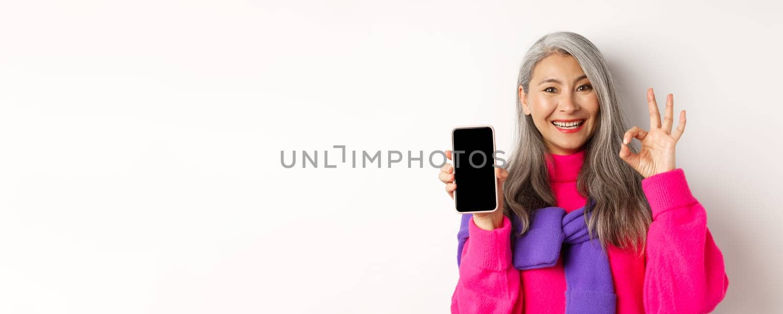 Online shopping. Fashionable asian senior woman showing blank smartphone screen and OK sign, smiling pleased, recommending app, white background.
