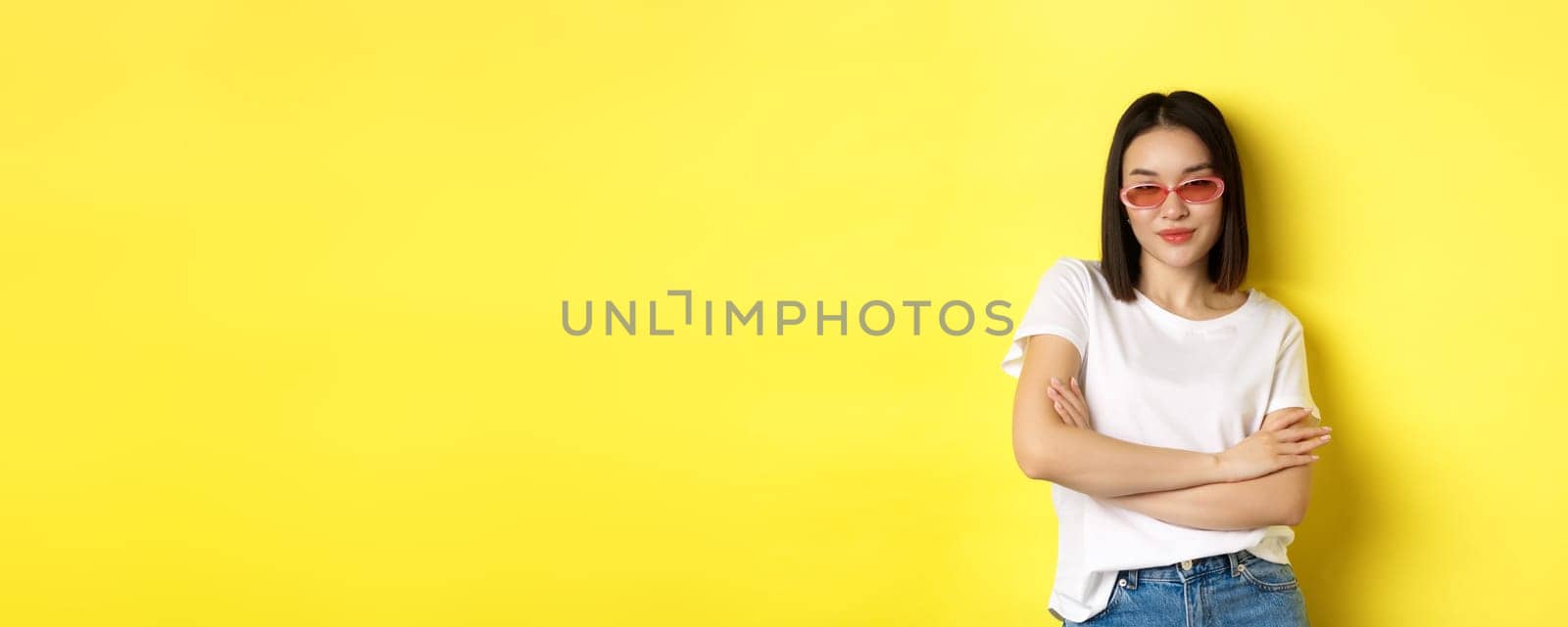 Fashion and lifestyle concept. Sassy and confident asian woman in trendy sunglasses looking self-assured at camera, standing over yellow background by Benzoix