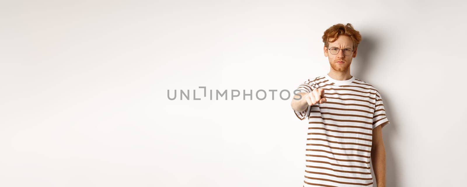 Angry and serious guy with red hair accusing you, frowning and pointing finger at camera, blame someone, standing over white background.