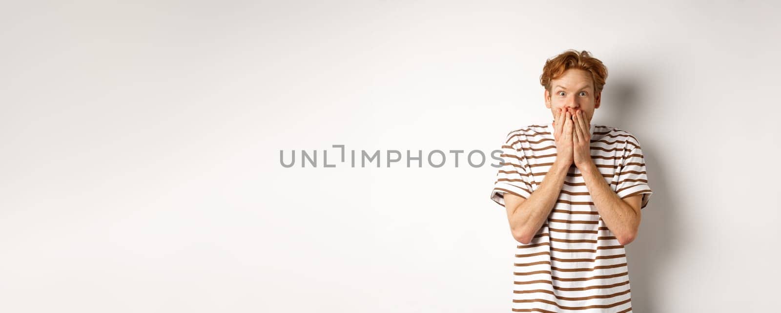 Surprised and excited man with curly red hair checking out special promo offer,staring fascinated and amazed, covering mouth with hands, standing over white background by Benzoix