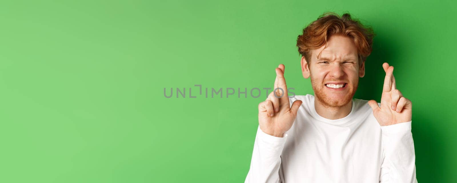 Hopeful caucasian man looking worried, waiting for important results and making wish with fingers crossed, standing over green background by Benzoix