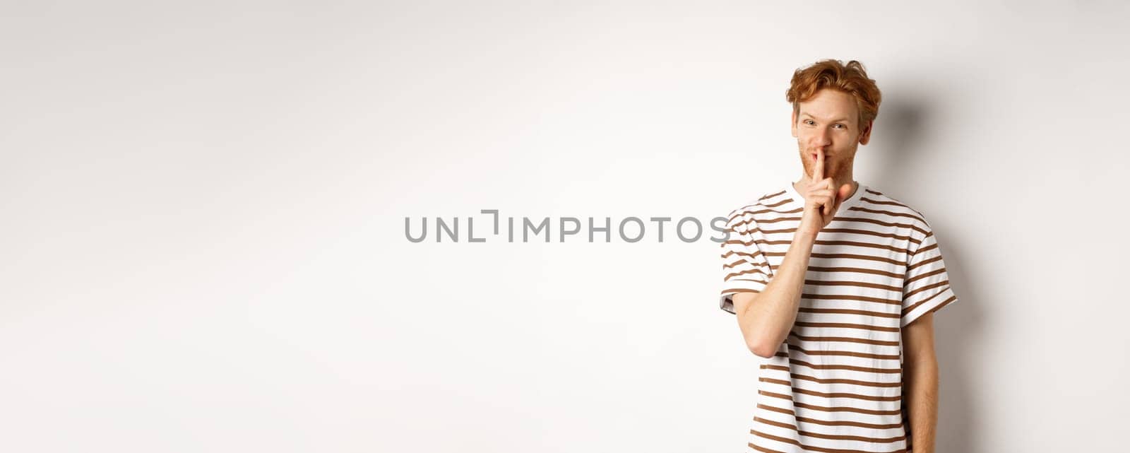 Young man student with red hair shushing, telling a secret and smiling, standing over white background by Benzoix