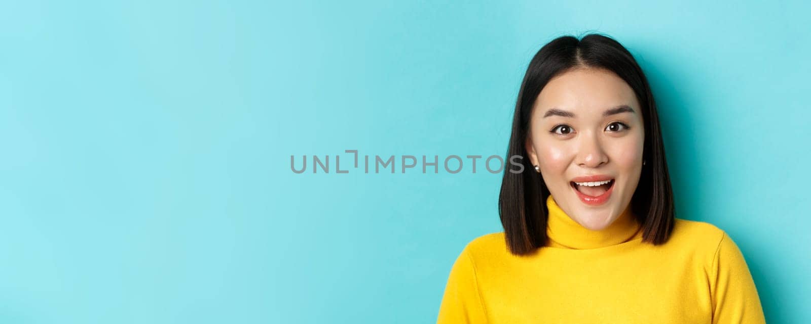 Beauty and skincare concept. Close up of surprised asian female raising eyebrows and look amused at camera, look at something interesting, standing over blue background.