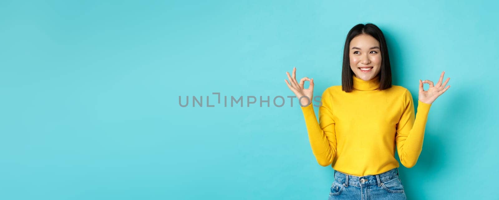 Beautiful japanese girl in yellow sweater, showing Ok signs and smiling, looking left and logo and recommending company, standing over blue background.