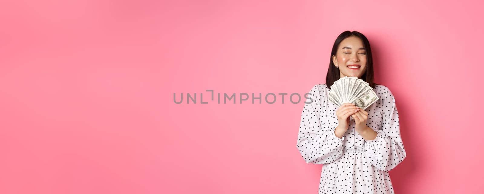 Shopping concept. Happy and satisfied asian woman winning prize money, showing dollars and rejoicing, standing over pink background.