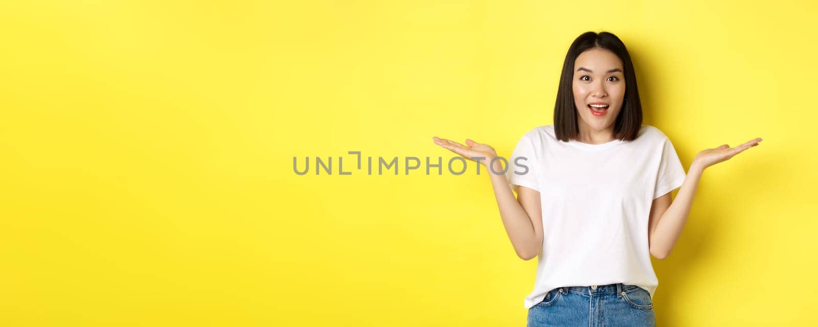 Beauty and fashion concept. Surprised asian girl spread hands sideways, holding on palms, pointing at two products, standing over yellow background.