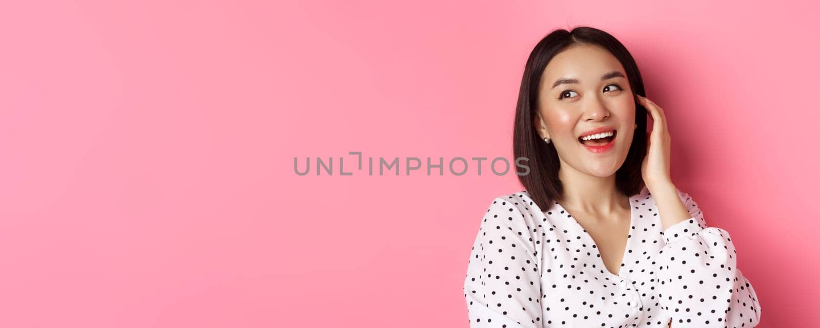 Close-up of flirty asian woman sighing, looking dreamy left and smiling, standing over pink background. Copy space