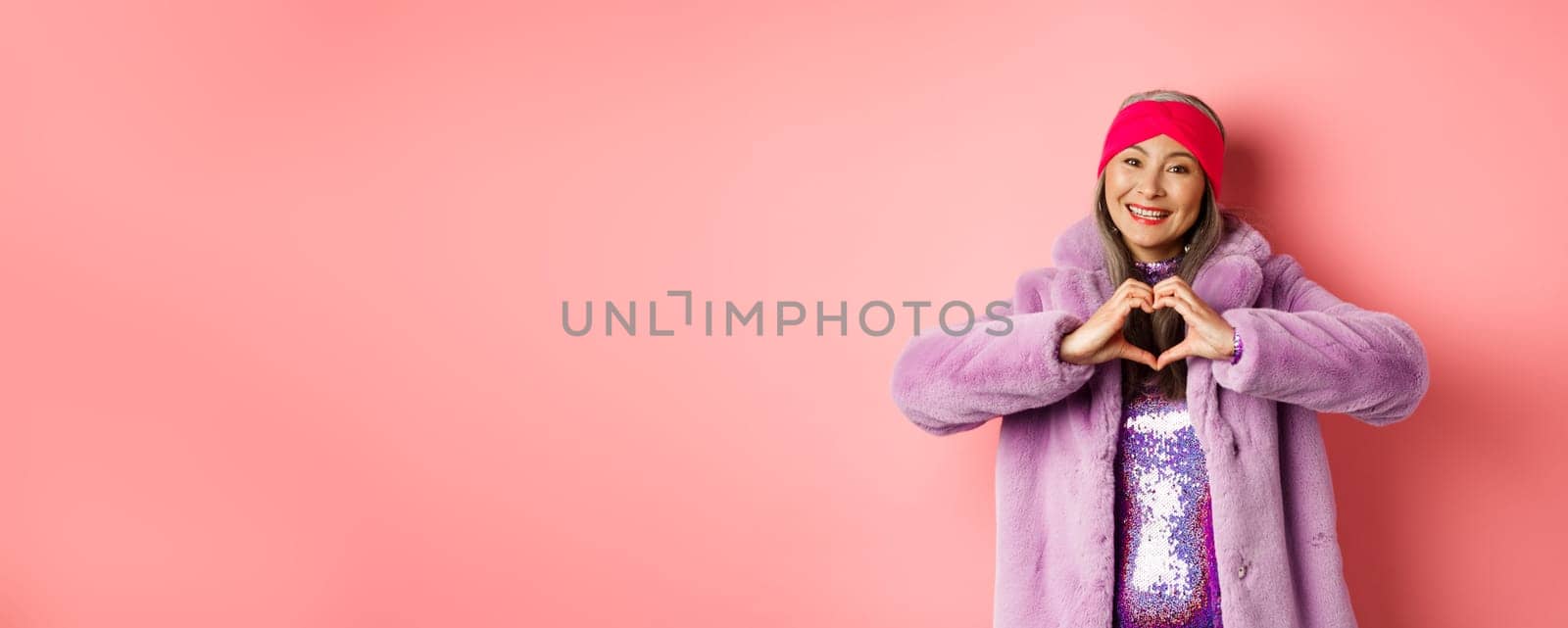 Romance and valentines day. Happy asian senior woman showing heart sign, I love you gesture, smiling and looking caring at camera, standing in faux fur coat, pink background.