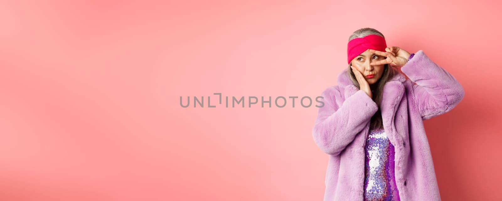 Fashion and shopping. Stylish and funky asian female model, showing peace signs and looking cute and silly, posing in purple faux fur coat over pink background.