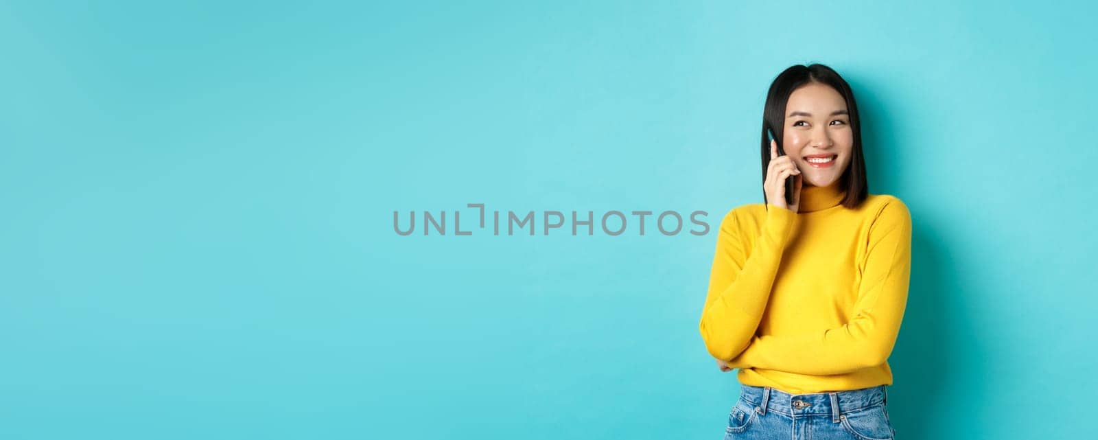 Attractive korean woman in yellow sweater, having conversation and smiling, talking on mobile phone, looking left at copy space, standing over blue background by Benzoix