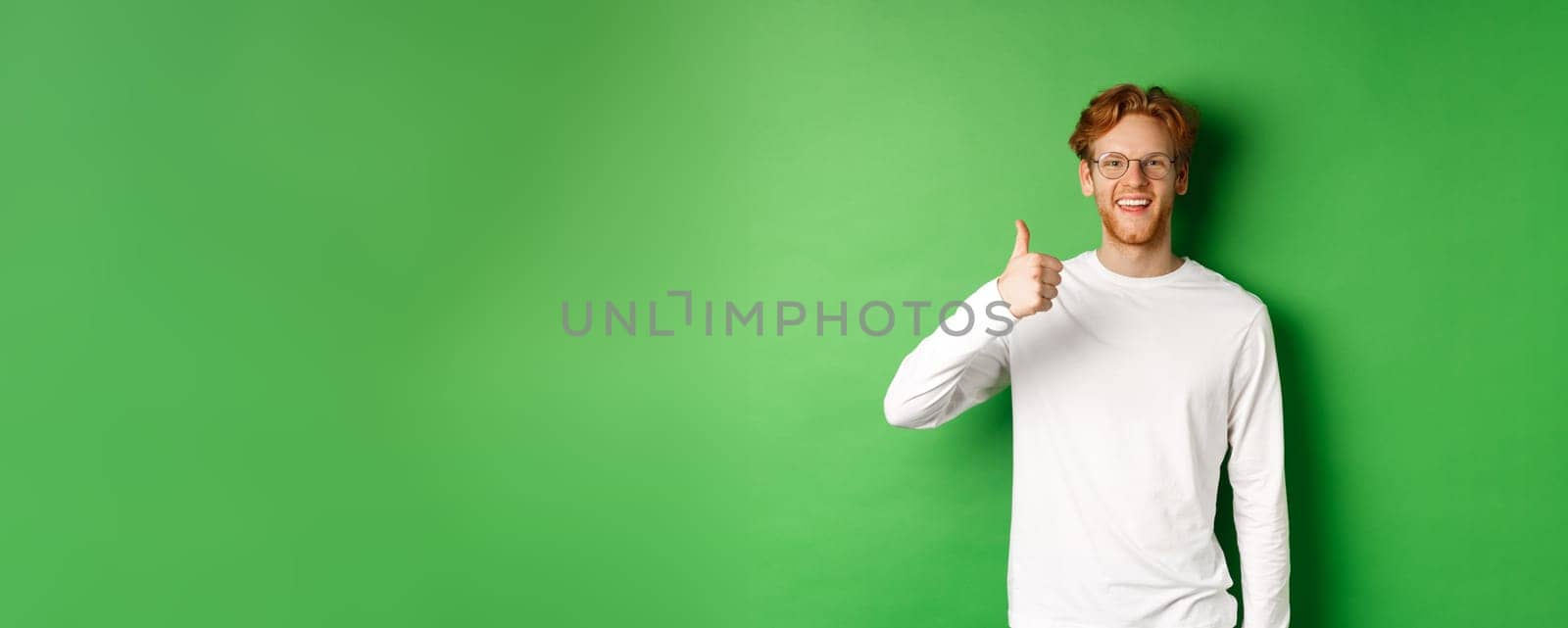 Handsome young man in glasses with red hair, showing thumbs-up and smiling satisfied, praising good eyewear store, standing over green background by Benzoix