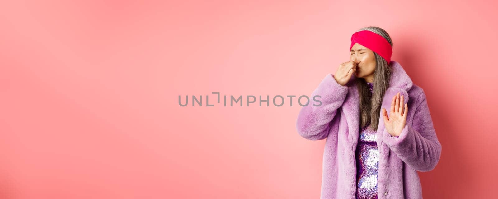 Disgusted asian mature woman in trendy purple winter coat and dress, shut nose and showing stop, refusal gesture, smell something disgusting, standing over pink background.
