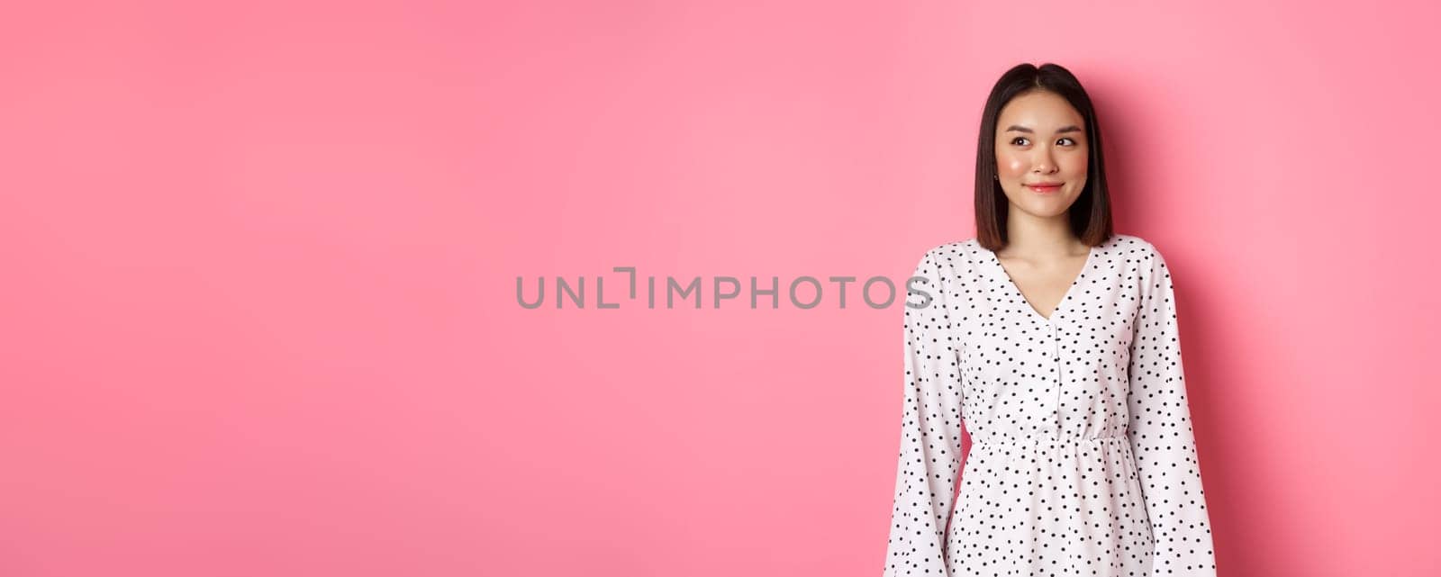 Cute asian woman smiling, looking left at copy space, standing on romantic pink background.