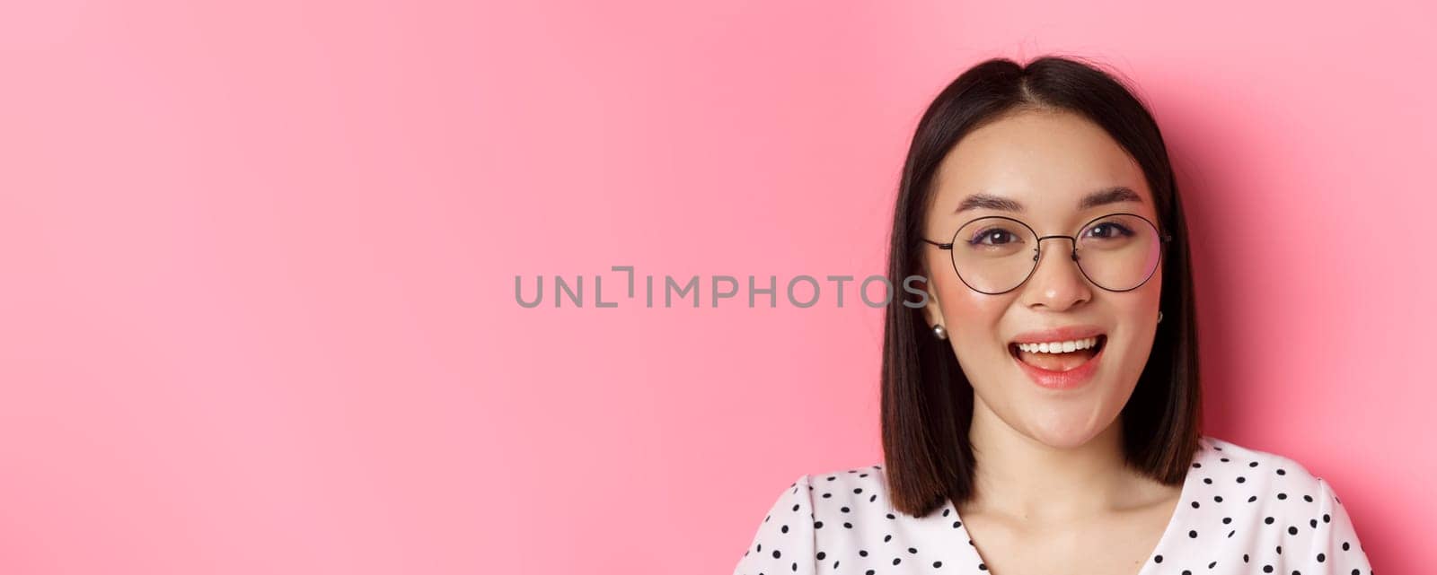 Beauty and lifestyle concept. Close-up of cute asian female model wearing trendy glasses, smiling happy at camera, standing on pink background by Benzoix