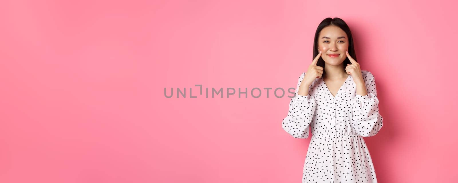Cute blushing asian girl poking cheeks, smiling happy at camera, wearing romantic white dress, standing over pink background.