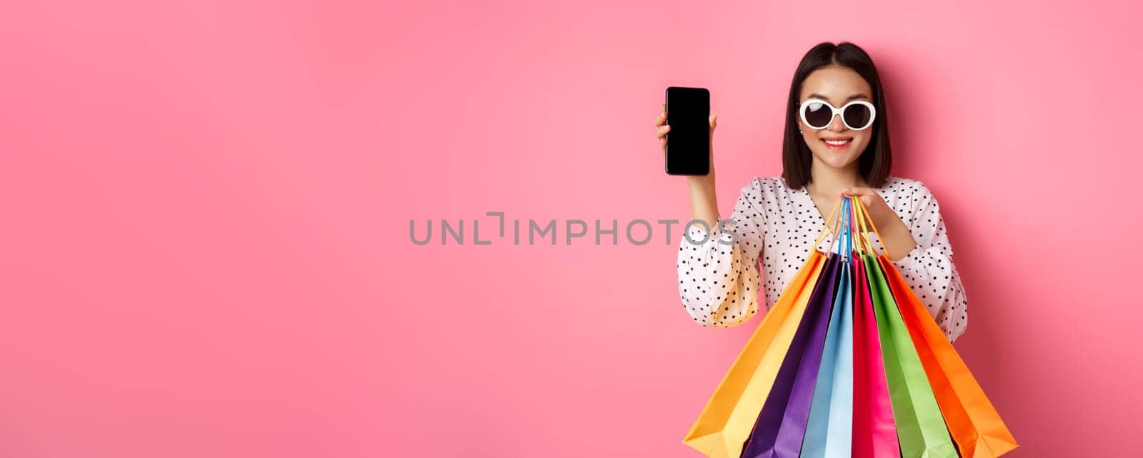 Attractive asian woman showing smartphone app and shopping bags, buying online via application, standing over pink background by Benzoix