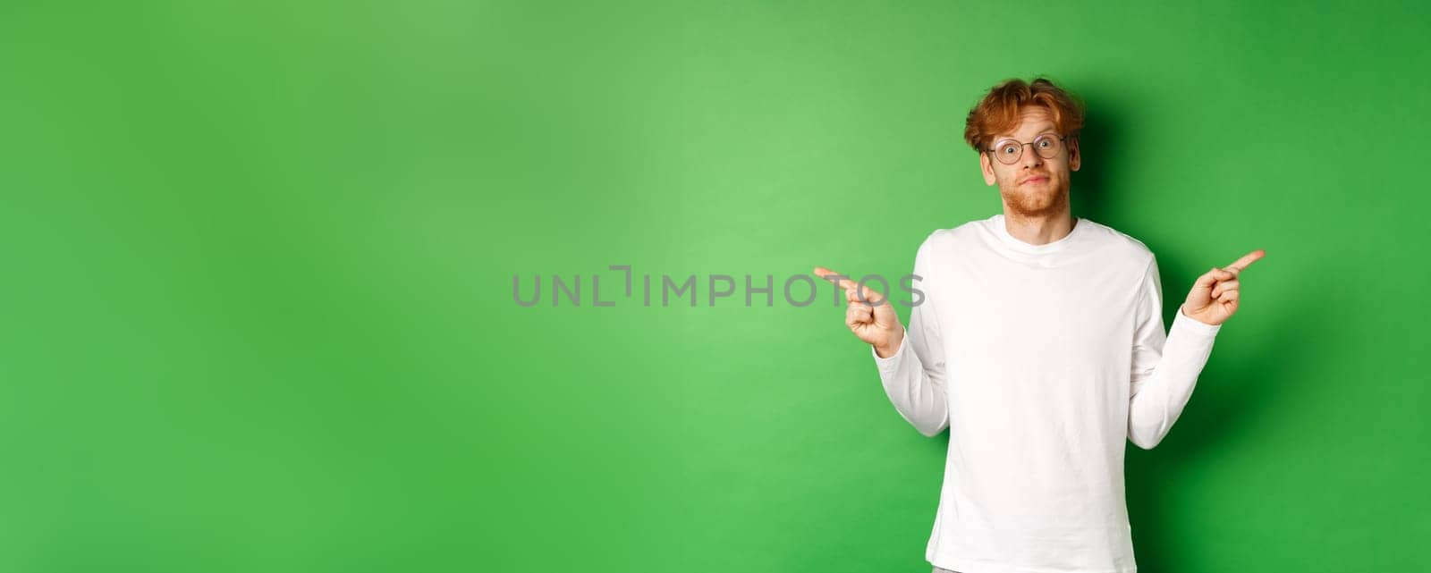 Indecisive funny guy with red hair pointing fingers sideways, staring confused at camera and showing two promo offers, standing over green background.