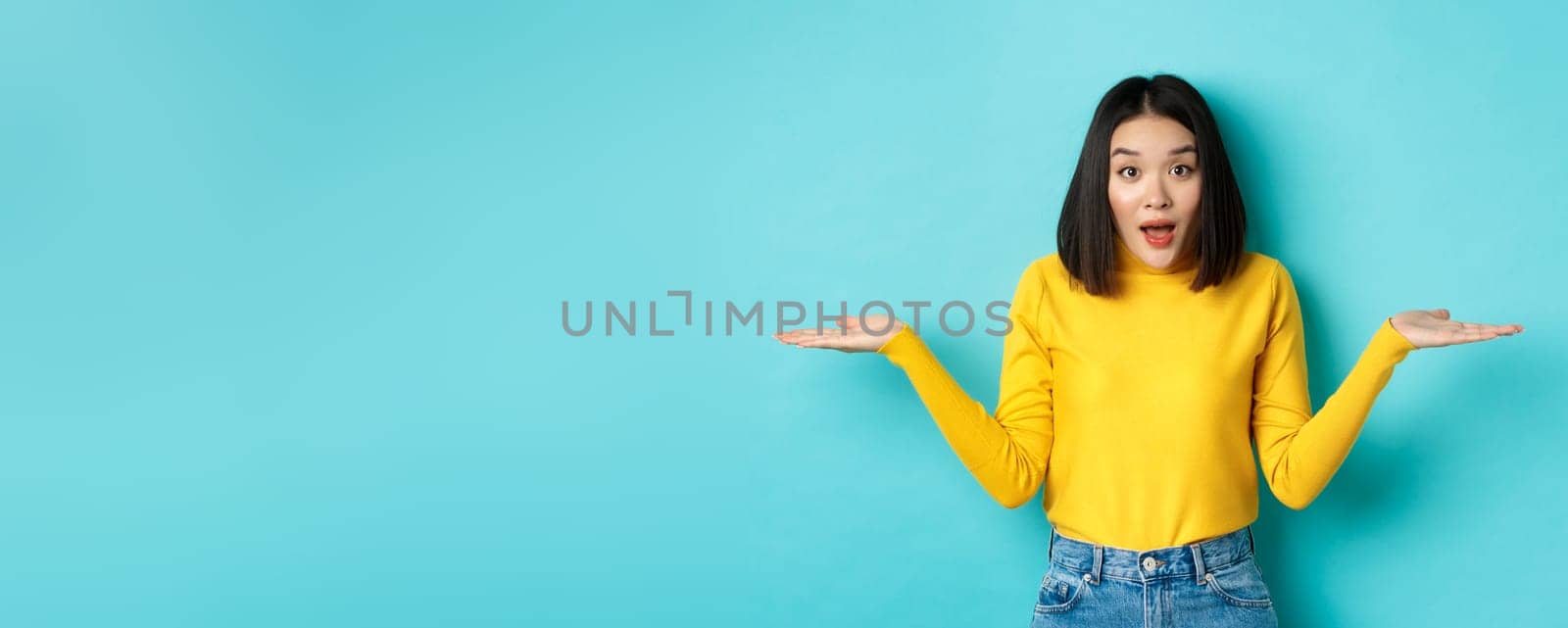 Image of indecisive asian woman shrugging shoulders, spread hands sideways and looking clueless at camera, standing confused against blue background.
