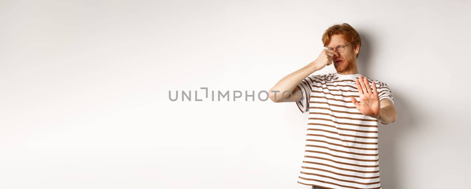Young man with red hair staring at camera and shut nose from bad smell, looking disgusted and refusing, white background.