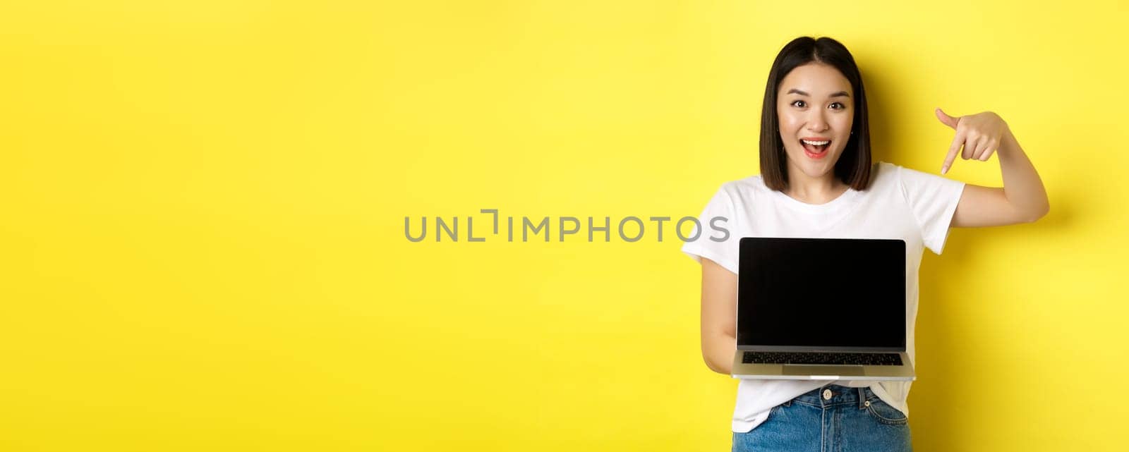 Young asian woman demonstrate online offer, showing laptop screen and smiling, standing over yellow background.
