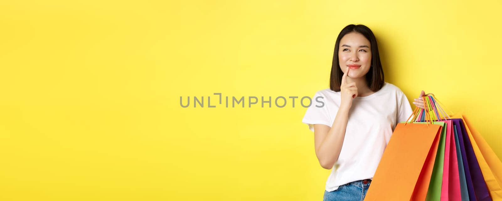 Pensive young woman smiling intrigued, showing shopping bags, thinking about buying something, standing over yellow background.