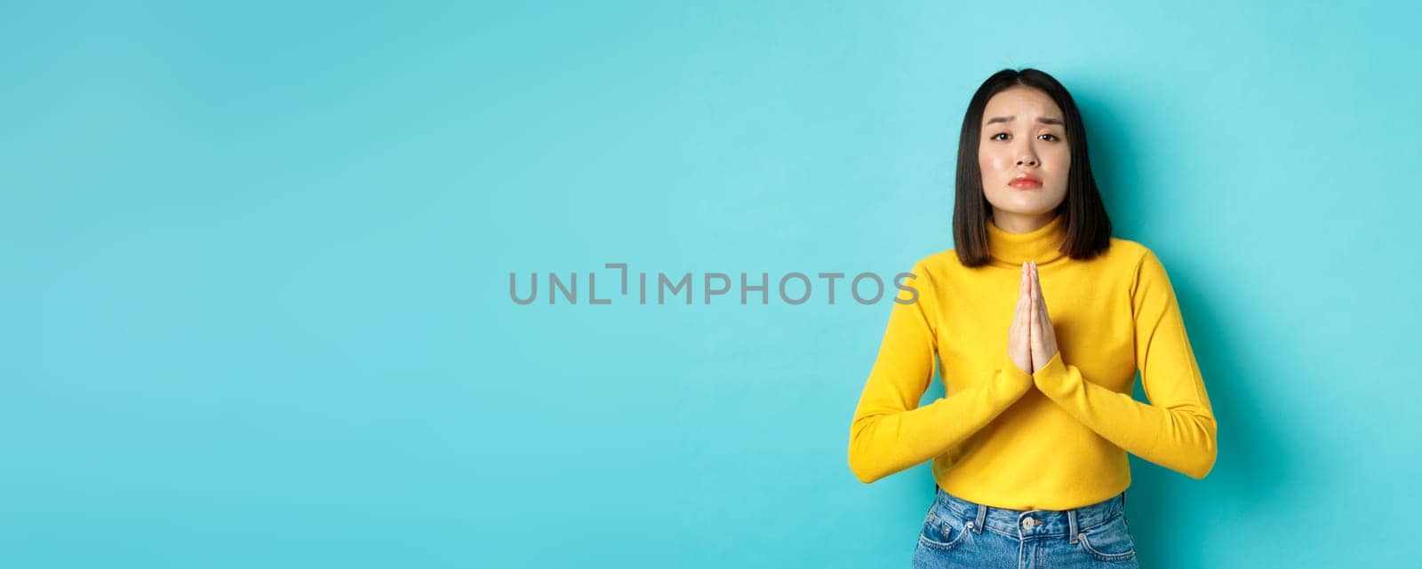 Young attractive asian girl begging for favour, need something and asking please, holding hands in pray, standing over blue background.