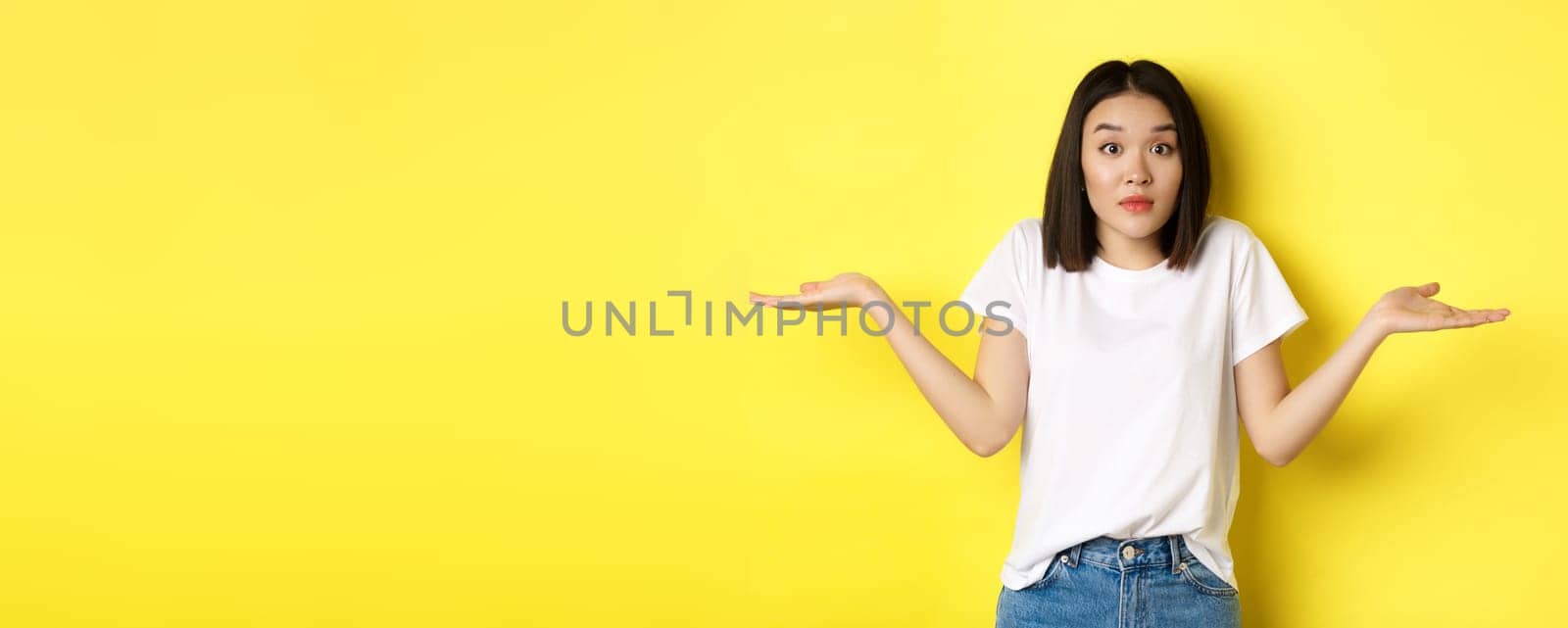 Clueless asian girl shrugging shoulders, spread out hands and looking confused, know nothing, standing over yellow background.