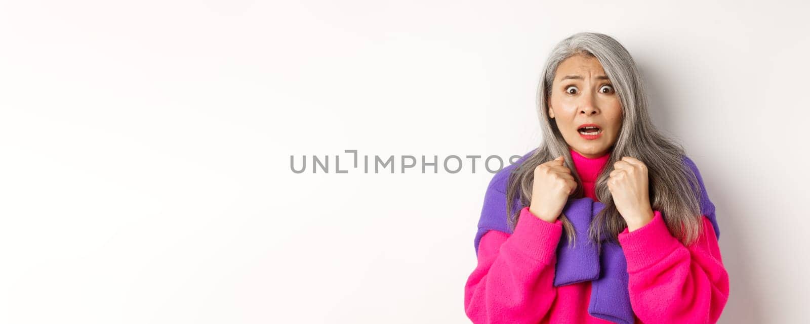 Close up of scared asian senior woman gasping, looking frightened and startled at camera, standing over white background.