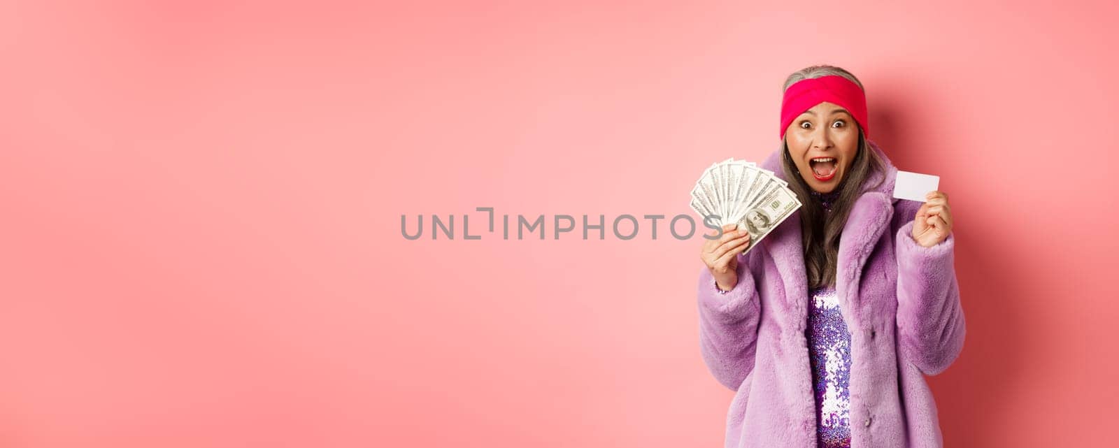 Shopping and fashion concept. Excited asian senior woman going buy something with money and plastic credit card, scream of joy and happiness, pink background.