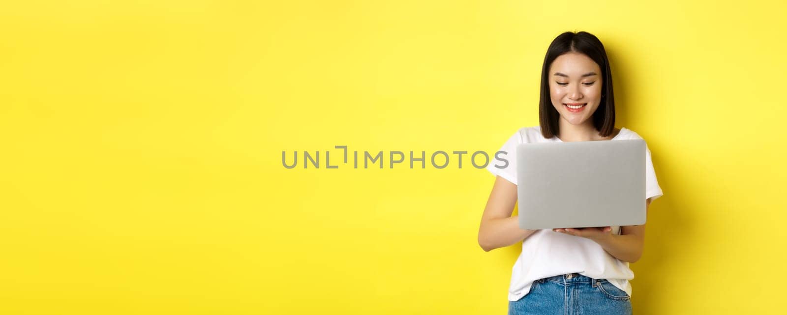 Beautiful asian woman working on laptop, looking happy at screen, standing over yellow background.