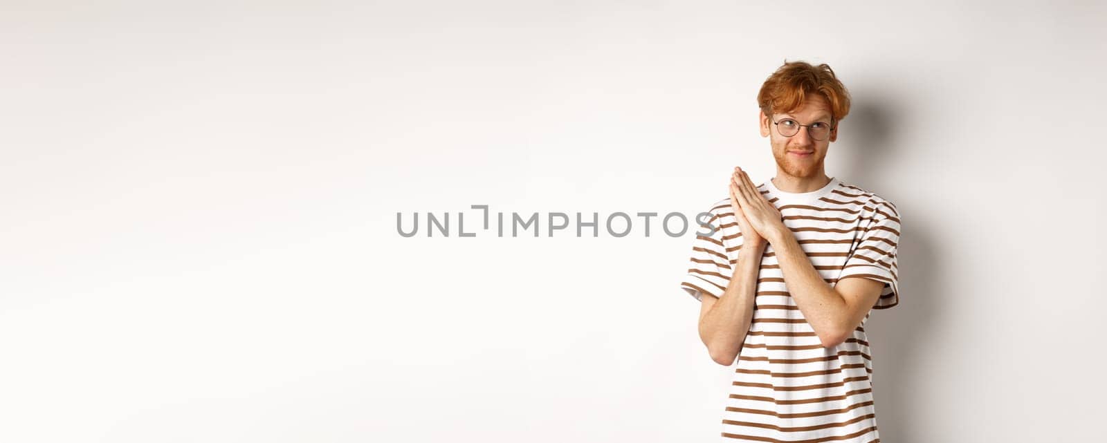 Thoughtful man with red hair rubbing hands and looking at upper right corner, having an idea, standing over white background.