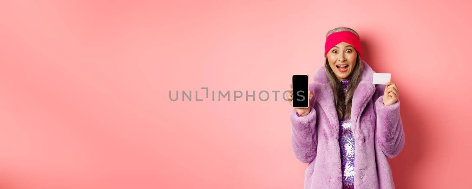 Special promotion. Excited asian senior woman showing blank smartphone screen and plastic credit card, checking out online offer, pink background by Benzoix