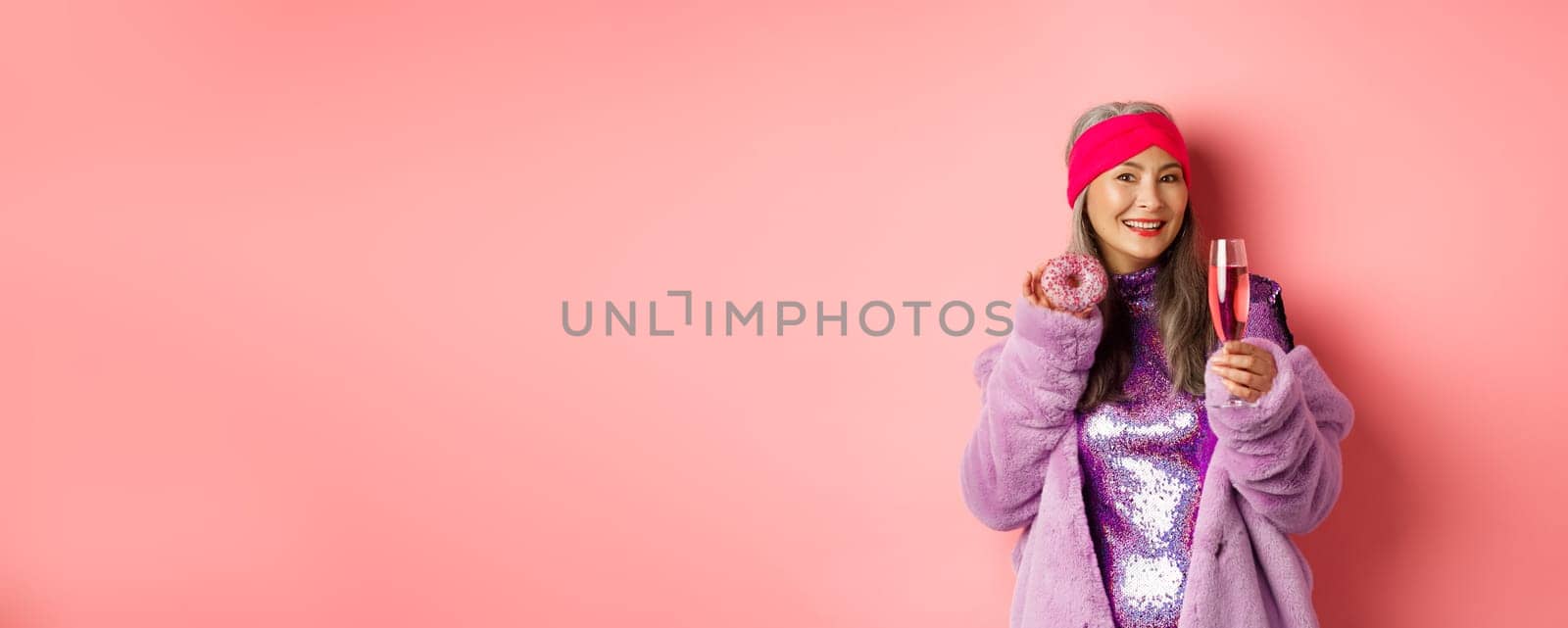 Elegant and stylish asian woman in purple faux fur coat eating donuts and drinking champagne, having fun at party, standing over pink background.