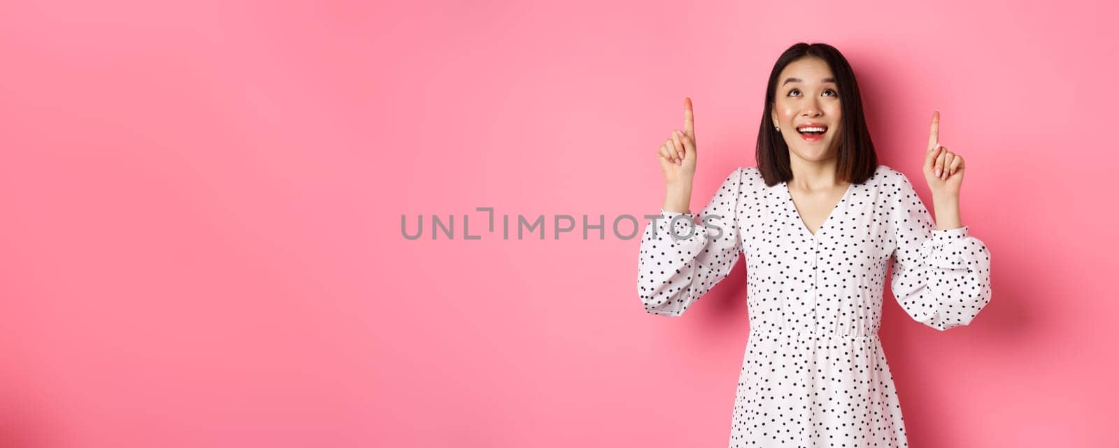 Amazed brunette girl in dress looking, pointing fingers up, showing logo with wondered smile, standing over pink background.