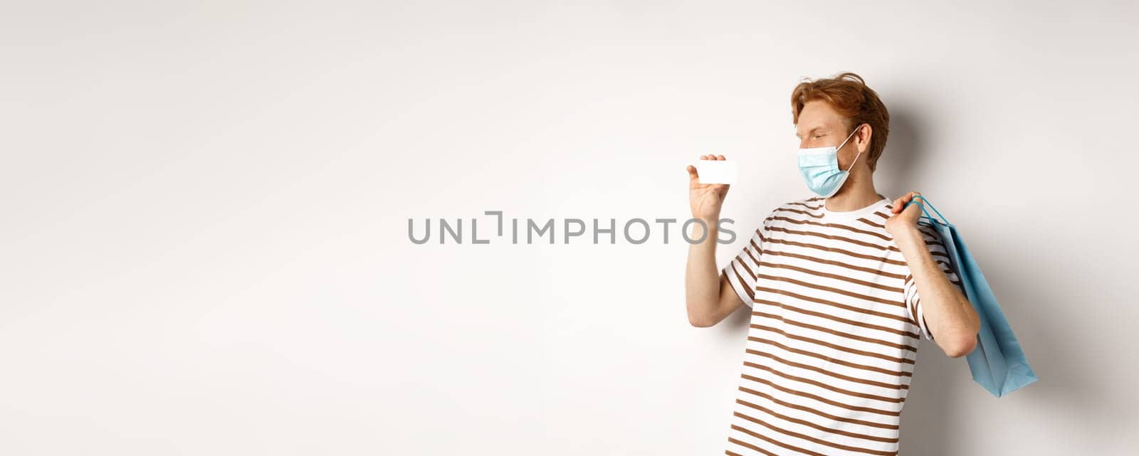 Concept of covid-19 and shopping. Happy young shopper in face mask holding paper bag and showing plastic credit card, buying with discounts, white background.