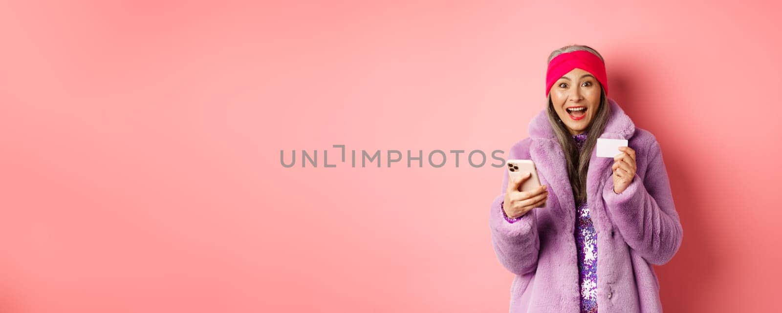 Online shopping and fashion concept. Happy asian female checking out internet promotions, holding credit card and smartphone, pink background by Benzoix