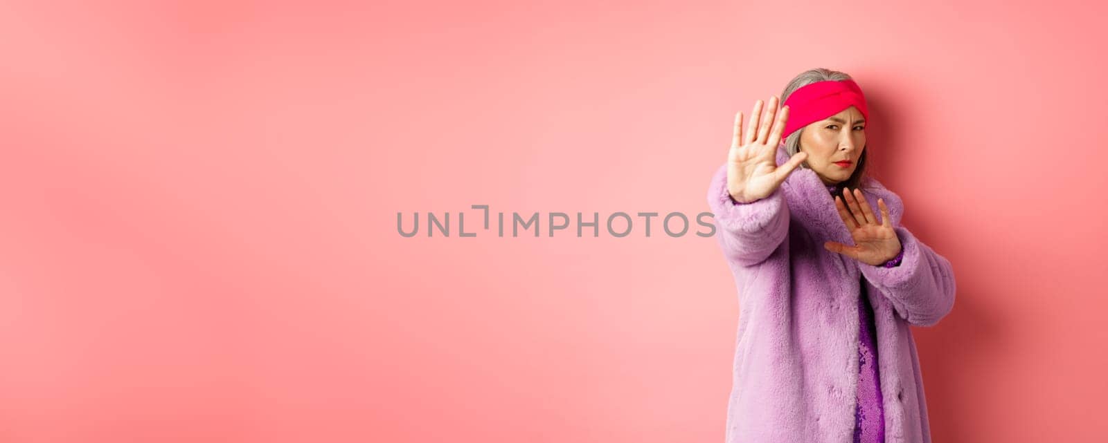 Asian senior female model in stylish purple winter coat standing in victim pose, extending hand in stop gesture and pleading for mercy, standing against pink background.