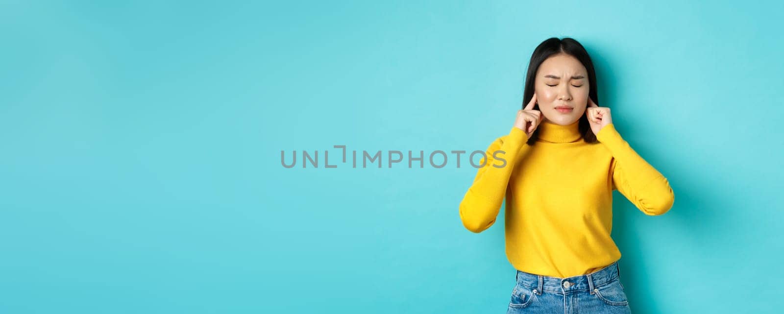 Tired and disappointed young asian woman unwilling to listen, shut ears with fingers and close eyes, standing in denial over blue background.