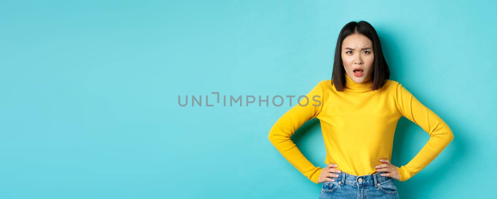 Portrait of angry and confused asian woman staring frustrated at camera, standing against blue background.