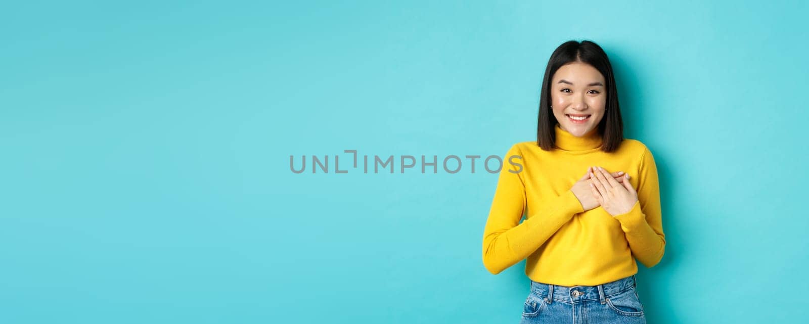 Image of beautiful asian woman holding hands on heart and smiling, thanking you, feeling grateful, standing over blue background.