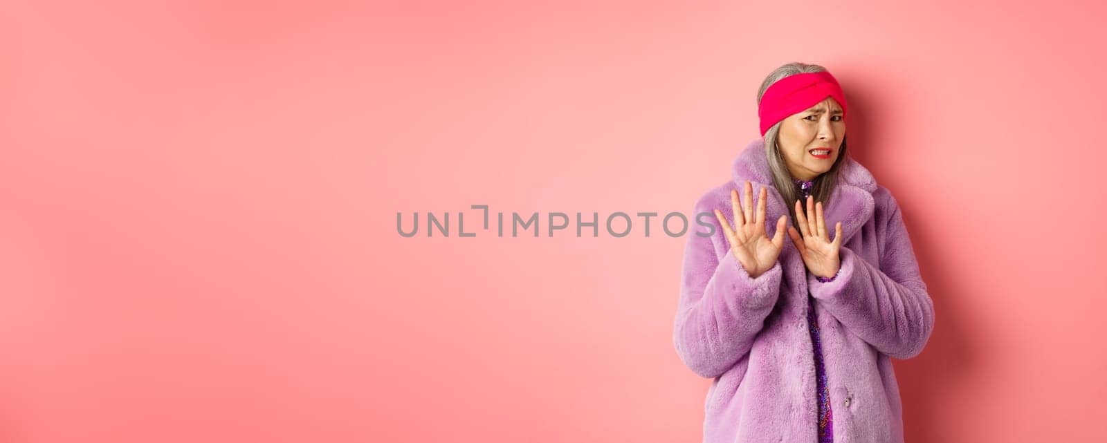 Portrait of stylish asian senior woman in purple winter coat, cringe from something disgusting, frowning and turn away awkward, showing stop gesture, refusing on pink background by Benzoix