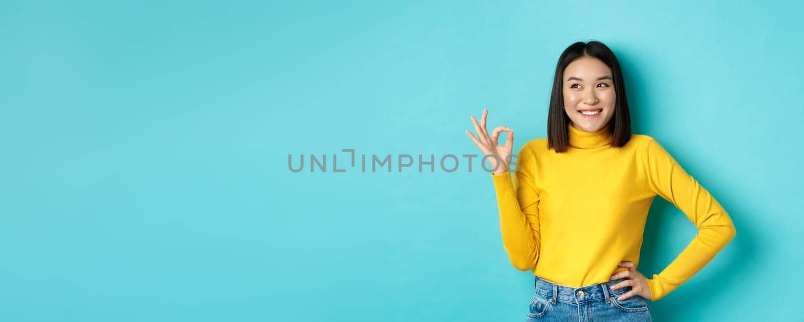 Impressed asian woman in yellow sweater, looking left amazed and showing OK sign in approval, standing over blue background by Benzoix