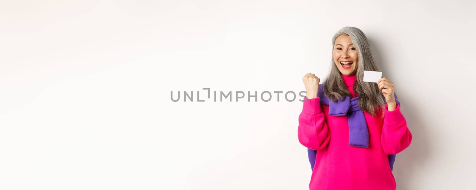 Shopping concept. Cheerful asian senior woman winning prize from bank, showing fist pump gesture and plastic credit card, standing happy over white background.