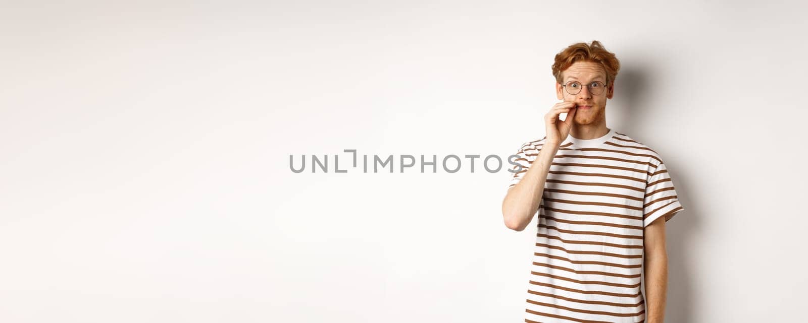 Young guy with ginger hair and glasses zipping mouth, showing lips seal gesture and smiling, keeping a secret, standing over white background.