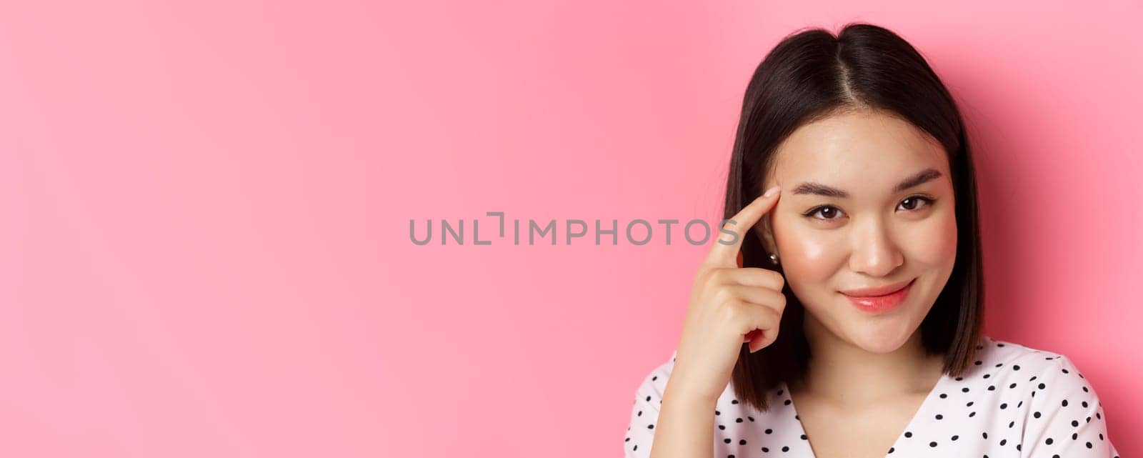 Beauty and skin care concept. Headshot of smart asian woman pointing at head and smiling sly, asking to think, standing over pink background.