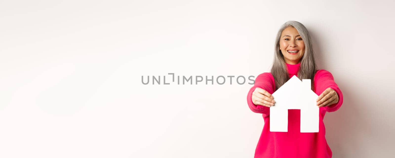 Real estate. Happy korean middle-aged woman with grey hair, showing paper house model and smiling, standing in pink sweater over white background by Benzoix