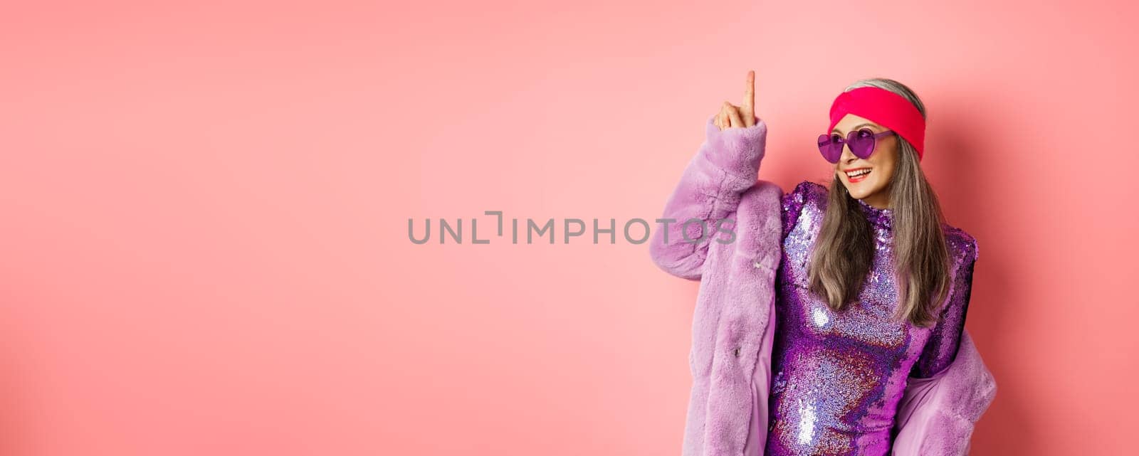 Asian old woman in fashionable glittering dress and purple faux fur coat dancing disco, pointing finger up and having fun, pink background by Benzoix