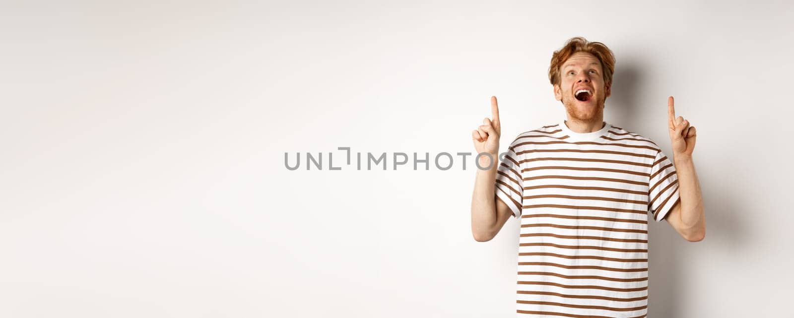 Fascinated redhead man looking with amazement and happiness, pointing fingers up, checking out something cool, standing over white background by Benzoix