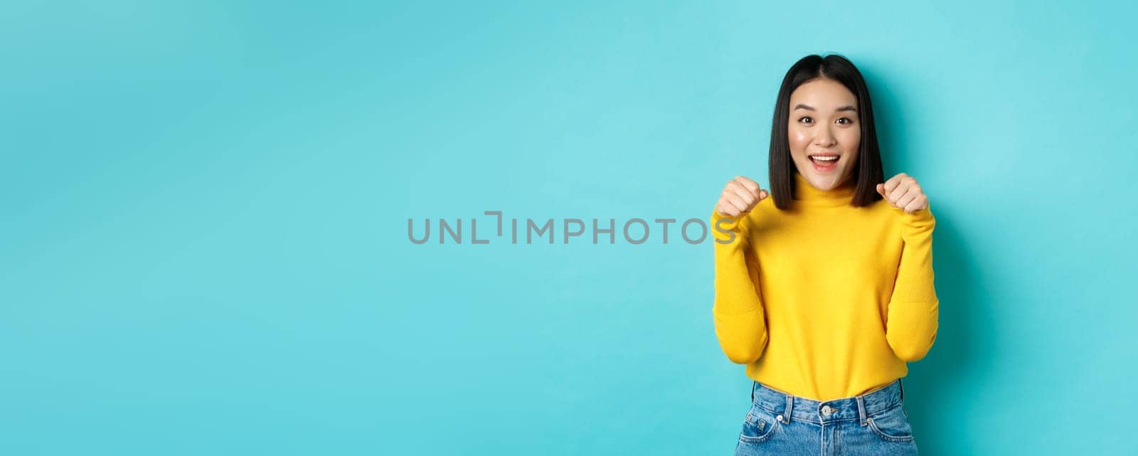 Beauty and fashion concept. Beautiful asian woman in yellow pullover raise hands as if holding logo, standing over blue background.