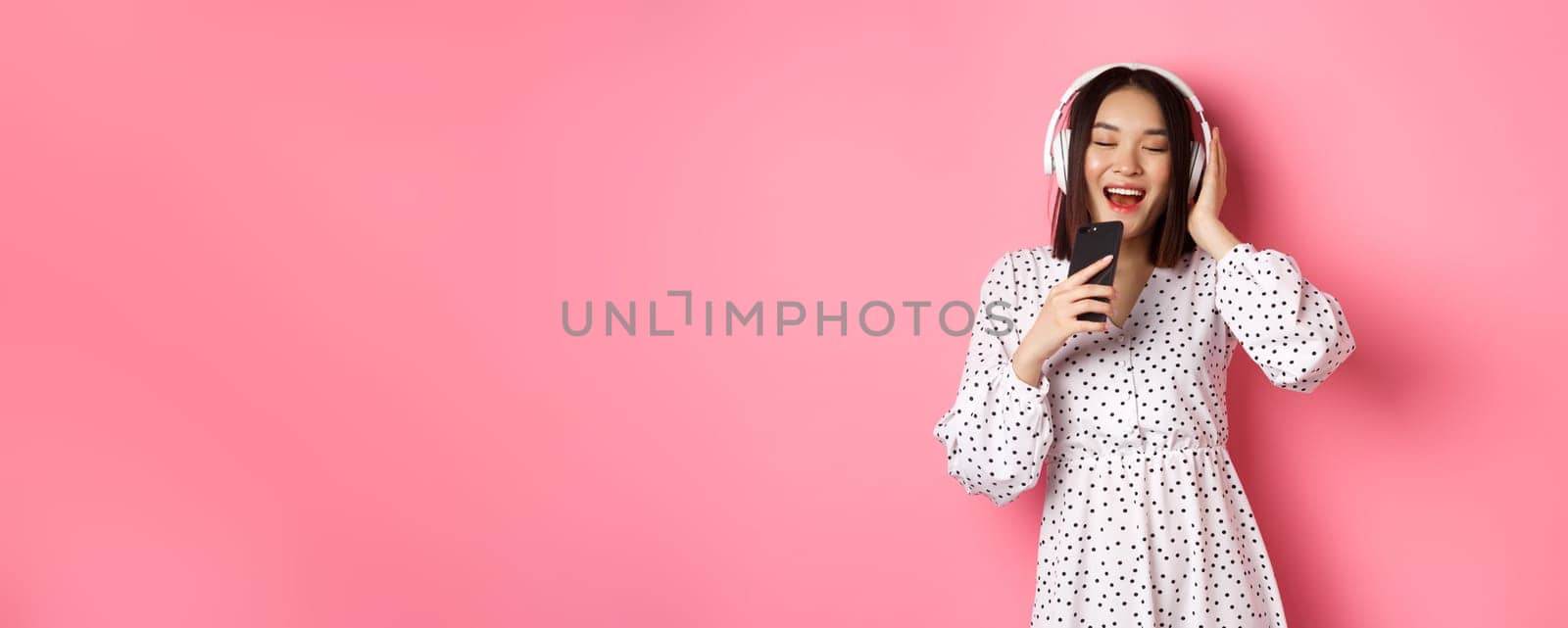 Cute asian woman playing karaoke app, singing in mobile phone and using headphones, standing in dress over pink background.
